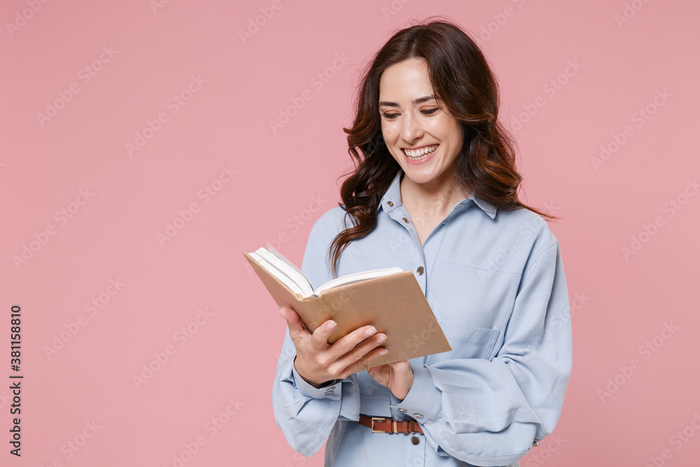 Smiling attractive beautiful pretty young brunette woman 20s wearing casual blue shirt dress posing standing holding in hands reading book isolated on pastel pink colour background, studio portrait. Smiling attractive beautiful pretty young brunette woman 20s wearing casual blue shirt dress posing standing holding in hands reading book isolated on pastel pink colour background, studio portrait.