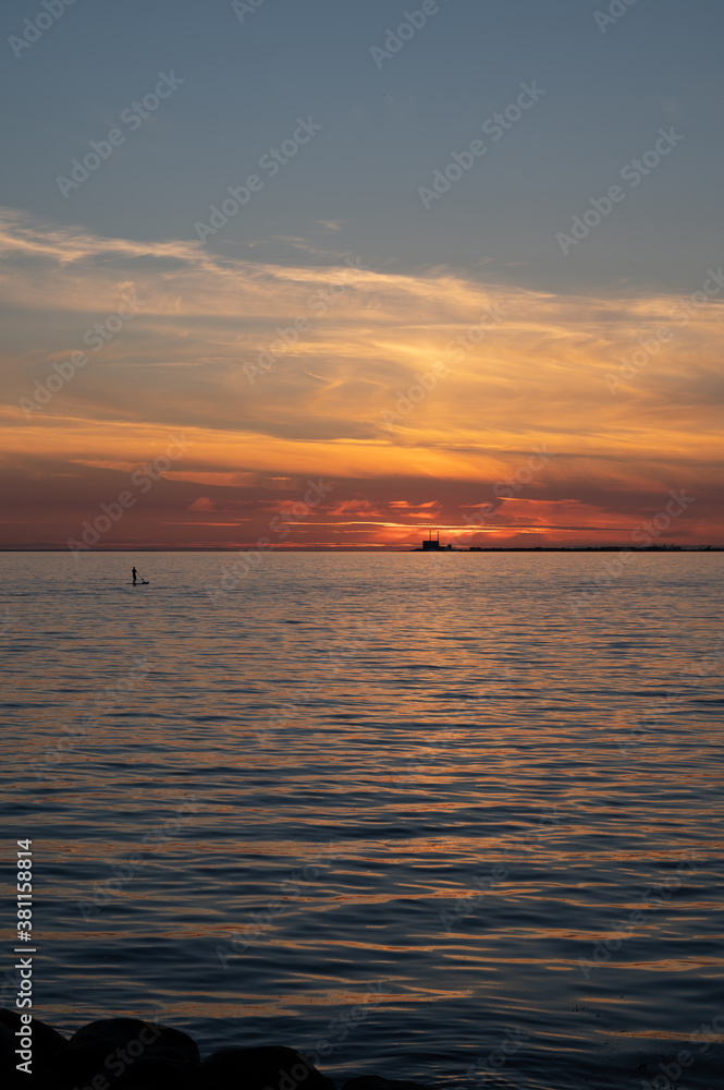 Obraz premium A man standing on a surf board on the calm bay water with the nuclear power plant Barsebäck visible on the horizon during summer in Sweden