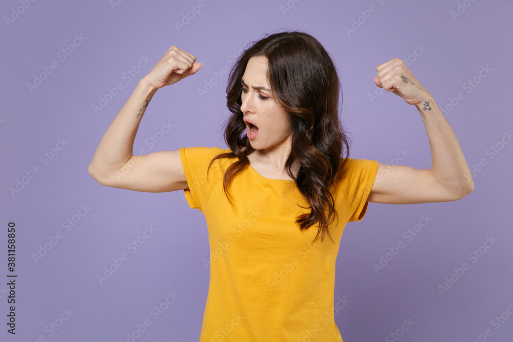 Shocked strong young brunette woman 20s wearing basic yellow t-shirt posing standing spreading hands showing biceps muscles looking aside isolated on pastel violet colour background, studio portrait. Shocked strong young brunette woman 20s wearing basic yellow t-shirt posing standing spreading hands showing biceps muscles looking aside isolated on pastel violet colour background, studio portrait.