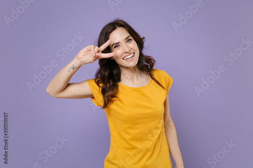 Smiling cheerful funny beautiful young brunette woman 20s wearing basic yellow t-shirt posing standing showing victory sign looking camera isolated on pastel violet colour background, studio portrait.