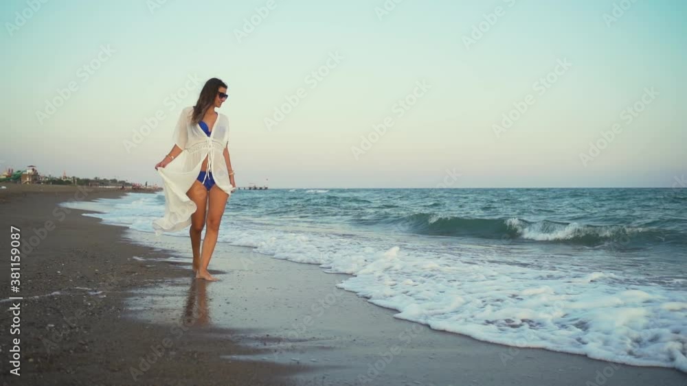 A young girl in sunglasses and a blue swimsuit walks along the sandy coast of the ocean. A white dress flutters in the wind. Summer beach vacation A young girl in sunglasses and a blue swimsuit walks along the sandy coast of the ocean. A white dress flutters in the wind. Summer beach vacation
