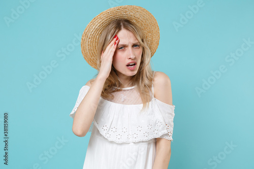 Preoccupied puzzled displeased sick young blonde woman 20s in white summer dress hat stand put hand on head having headache looking camera isolated on blue turquoise colour background studio portrait. © ViDi Studio
