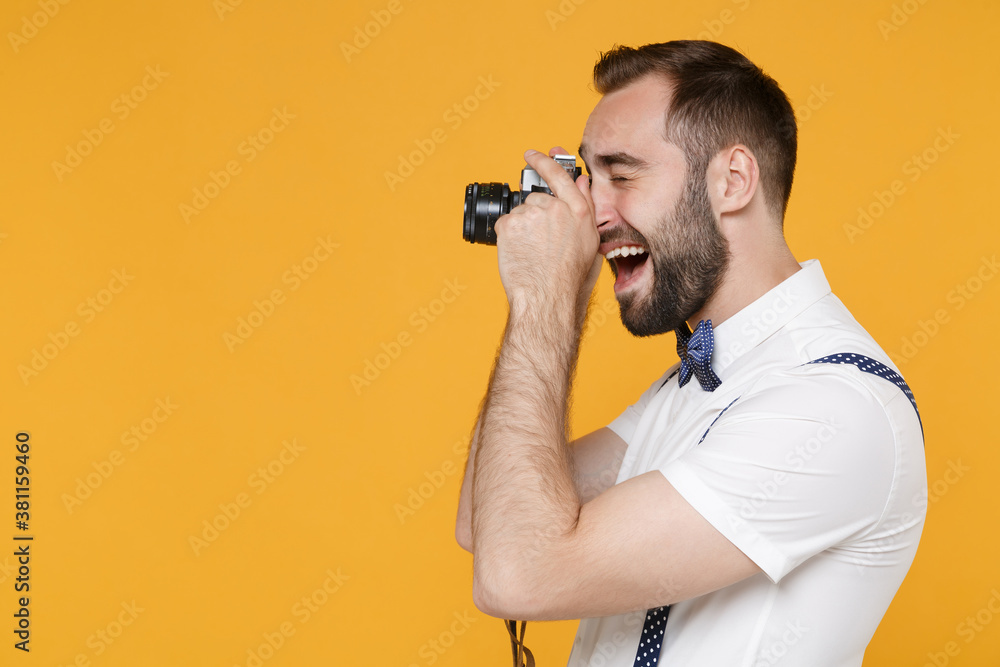 Side view of funny young bearded man 20s wearing white shirt bow-tie suspender posing taking picture with retro vintage photo camera isolated on bright yellow color wall background studio portrait.