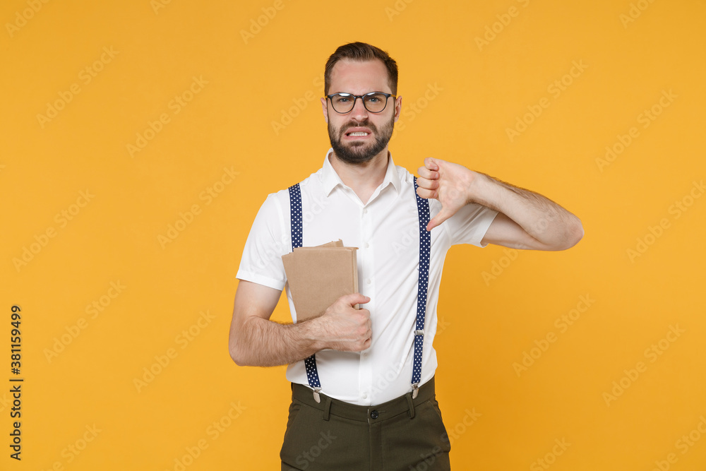 Displeased young bearded man 20s wearing white shirt suspender eyeglasses posing standing hold in hands books looking camera showing thumb down isolated on yellow color background, studio portrait. Displeased young bearded man 20s wearing white shirt suspender eyeglasses posing standing hold in hands books looking camera showing thumb down isolated on yellow color background, studio portrait.