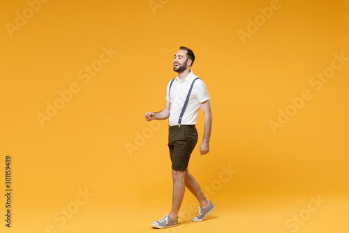 Full length side view portrait of cheerful funny young bearded man 20s wearing white shirt suspender shorts posing walking going looking aside isolated on bright yellow color wall background studio.