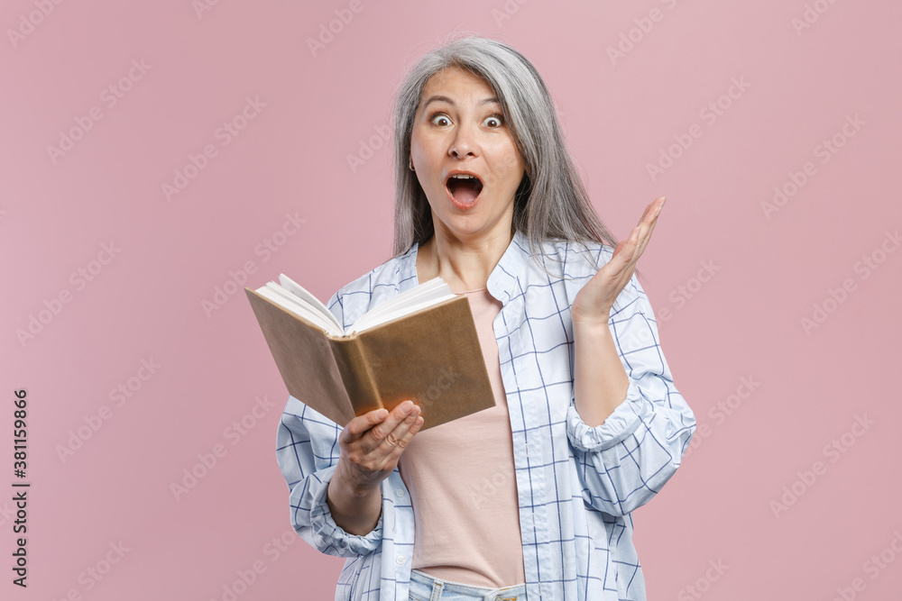 Shocked gray-haired asian woman wearing basic white checkered shirt standing holding in hands reading book spreding hands looking camera isolated on pastel pink colour background, studio portrait. Shocked gray-haired asian woman wearing basic white checkered shirt standing holding in hands reading book spreding hands looking camera isolated on pastel pink colour background, studio portrait.
