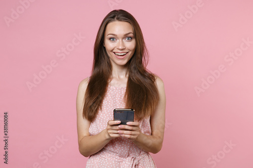 Smiling funny young brunette woman 20s wearing pink summer dotted dress posing using mobile cell phone typing sms message looking camera isolated on pastel pink color wall background studio portrait.