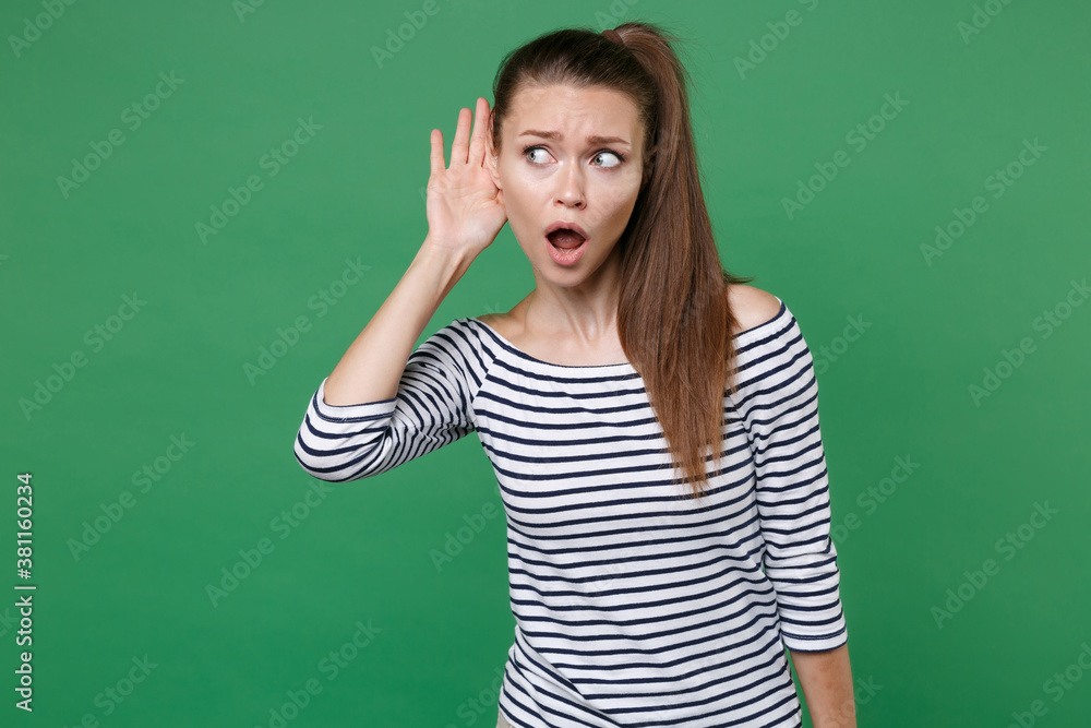 Shocked curious pretty young brunette woman 20s wearing striped casual clothes posing try to hear you overhear listening intently looking aside isolated on green color background, studio portrait.
