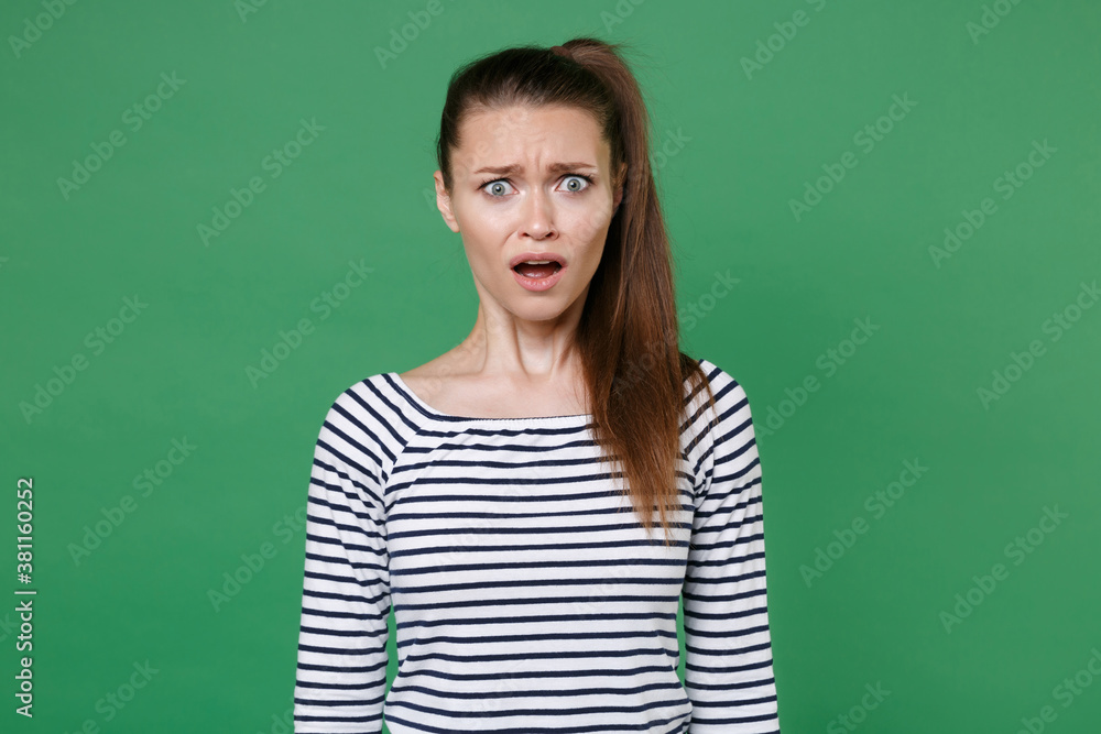 Shocked worried perplexed disgusted young brunette woman 20s wearing striped casual clothes posing standing keeping mouth open looking camera isolated on green color wall background, studio portrait. Shocked worried perplexed disgusted young brunette woman 20s wearing striped casual clothes posing standing keeping mouth open looking camera isolated on green color wall background, studio portrait.