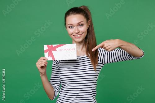 Smiling pretty beautiful young brunette woman 20s in striped casual clothes posing standing pointing index finger on gift certificate looking camera isolated on green color background studio portrait.