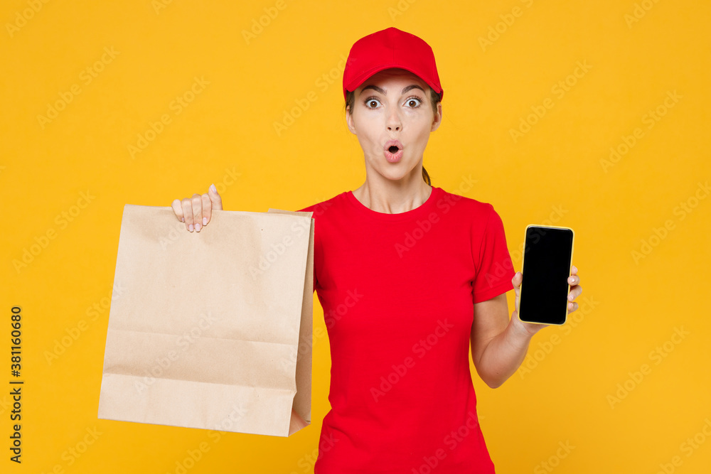 Delivery employee woman in red cap blank t-shirt uniform work courier service shop restaurant to home office hold mobile phone brown craft paper takeaway food bag mockup isolated on yellow background.