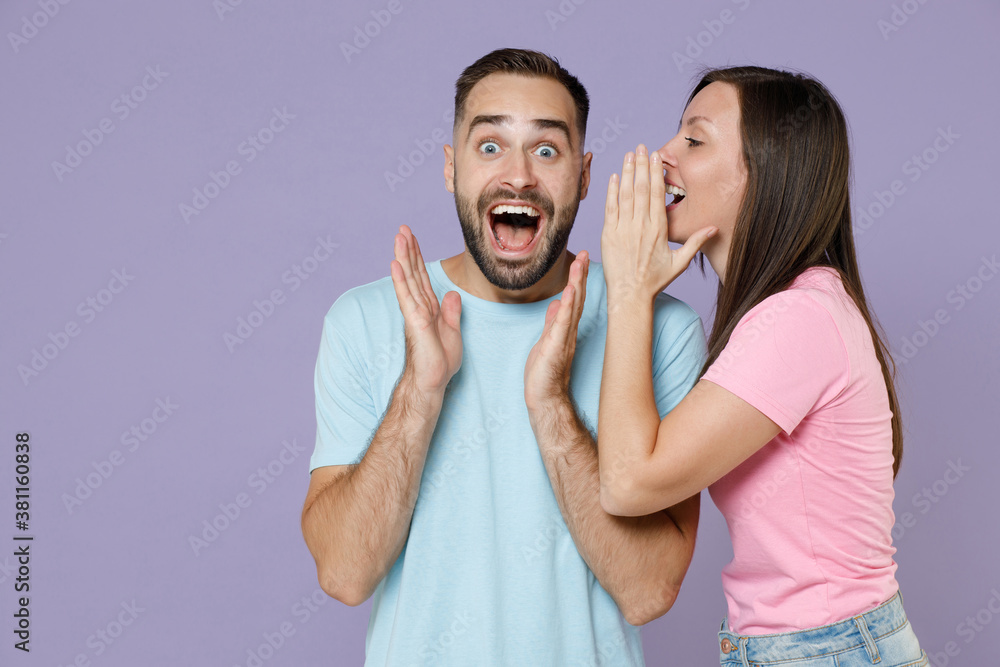 Surprised shocked young couple two friends man woman wearing blue pink blank design t-shirts standing whispering secret behind hand, sharing news isolated on pastel violet background studio portrait. Surprised shocked young couple two friends man woman wearing blue pink blank design t-shirts standing whispering secret behind hand, sharing news isolated on pastel violet background studio portrait.