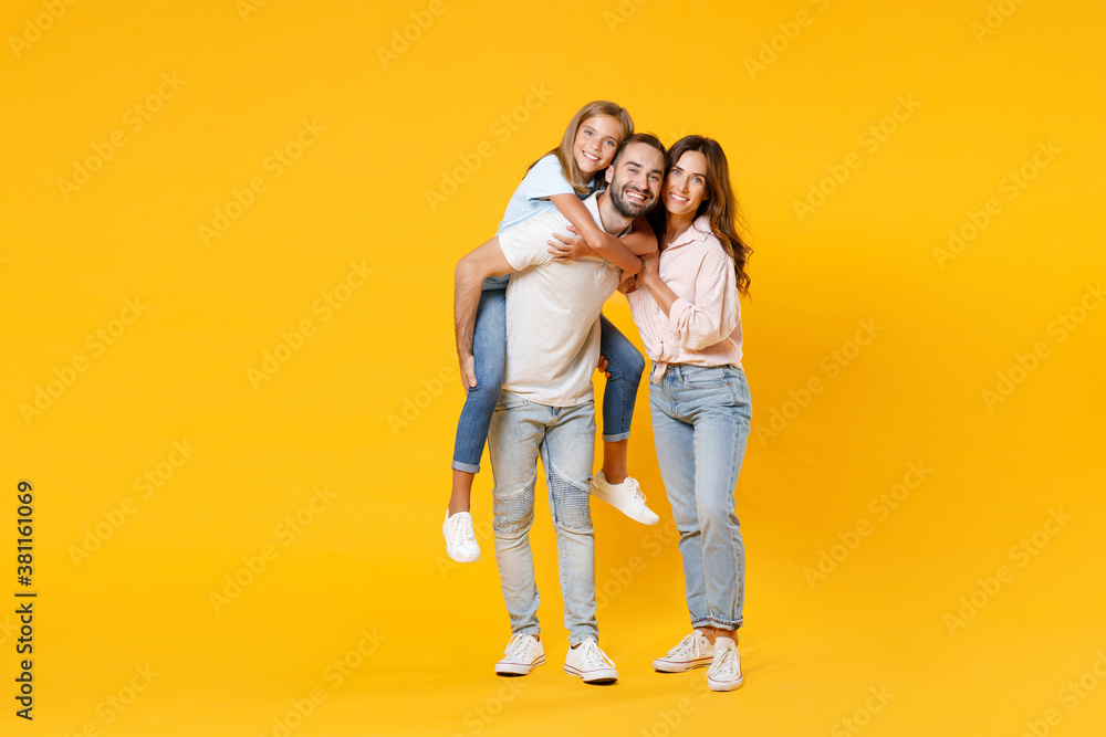 Full length portrait of smiling funny young parents mom dad with child kid daughter teen girl in t-shirts giving piggyback ride to joyful sit on back isolated on yellow background. Family day concept.