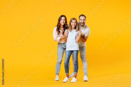 Full length portrait excited cheerful young parents mom dad with child kid daughter teen girl in t-shirts hugging looking camera isolated on yellow background. Family day parenthood childhood concept.