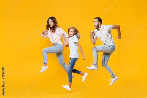 Full length portrait of cheerful funny young parents mom dad with child kid daughter teen girl in basic t-shirts jumping like running isolated on yellow background studio portrait. Family day concept. © ViDi Studio