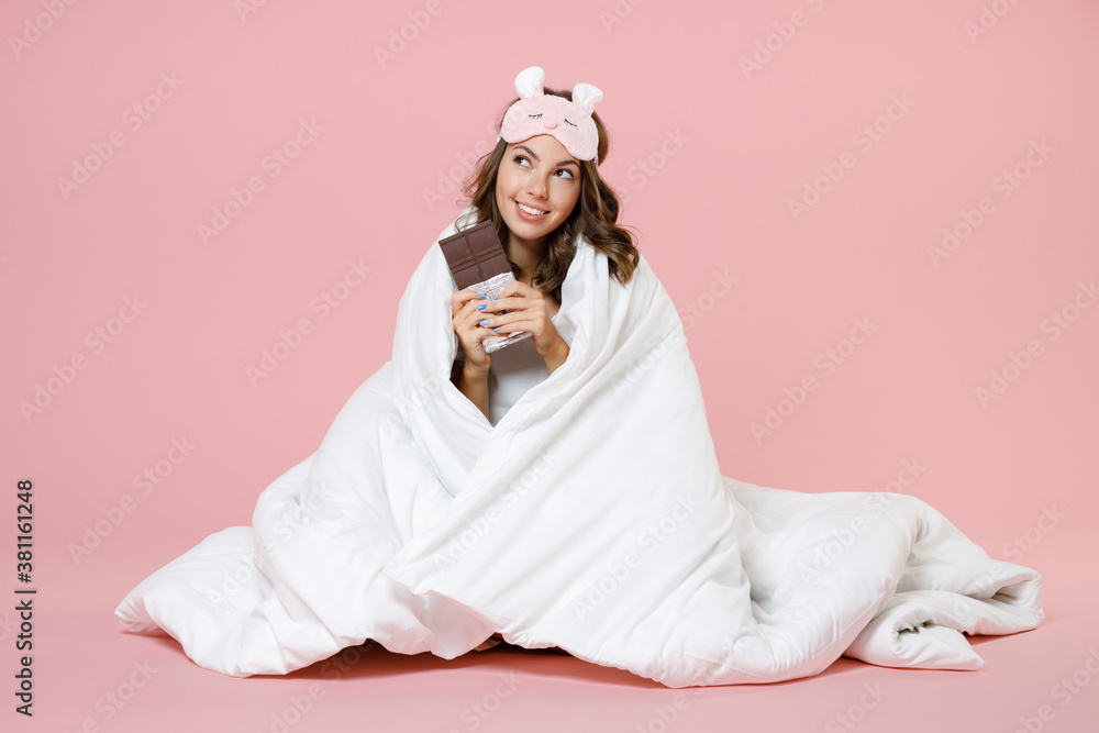 Smiling pensive young woman in pajamas home wear sleep mask sit hold in hands chocolate bar while resting at home isolated on pastel pink background studio portrait. Relax good mood lifestyle concept. Smiling pensive young woman in pajamas home wear sleep mask sit hold in hands chocolate bar while resting at home isolated on pastel pink background studio portrait. Relax good mood lifestyle concept.