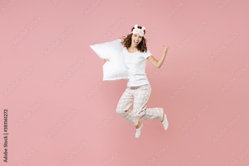 Full length portrait of happy young woman in white pajamas home wear jumping hold pillow doing winner gesture rest at home isolated on pastel pink background studio. Relax good mood lifestyle concept. - obrazy, fototapety, plakaty 