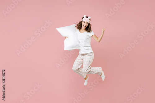 Full length portrait of happy young woman in white pajamas home wear jumping hold pillow doing winner gesture rest at home isolated on pastel pink background studio. Relax good mood lifestyle concept.