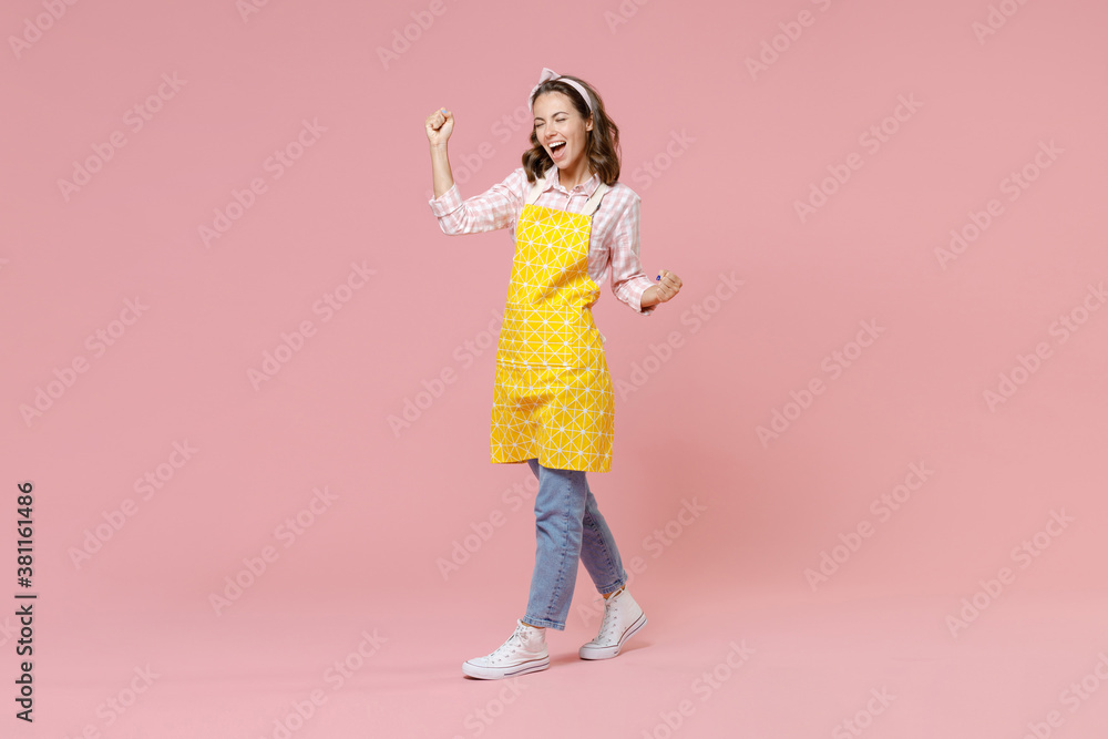Full length portrait joyful young woman housewife 20s in yellow apron doing winner gesture clenching fists keeping eyes closed isolated on pastel pink colour background studio. Housekeeping concept. Full length portrait joyful young woman housewife 20s in yellow apron doing winner gesture clenching fists keeping eyes closed isolated on pastel pink colour background studio. Housekeeping concept.