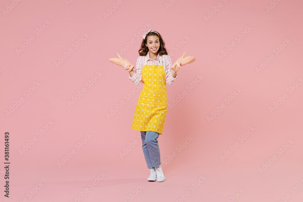 Full length portrait of smiling young brunette woman housewife in yellow apron gloves potholders spreading hands doing housework isolated on pastel pink colour background studio. Housekeeping concept. Full length portrait of smiling young brunette woman housewife in yellow apron gloves potholders spreading hands doing housework isolated on pastel pink colour background studio. Housekeeping concept.