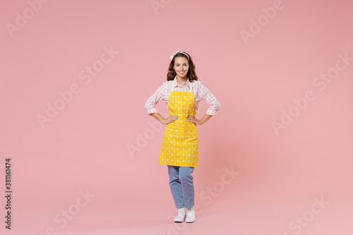 Full length portrait of smiling young brunette woman housewife wearing yellow apron standing with arms akimbo on waist doing housework isolated on pastel pink background studio. Housekeeping concept.