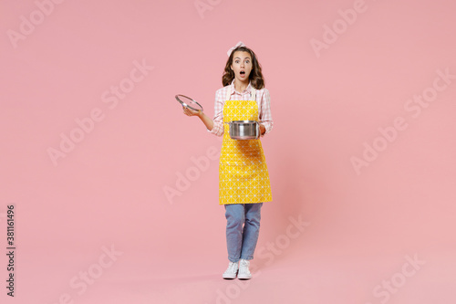 Full length portrait of shocked young brunette woman housewife 20s in yellow apron hold saucepan with lid while doing housework isolated on pastel pink colour background studio. Housekeeping concept.
