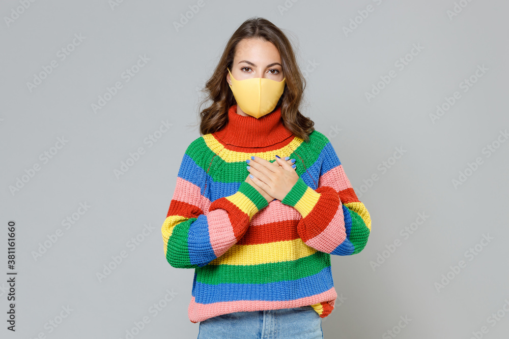 Young woman in colorful sweater face mask safe from coronavirus virus covid-19 during pandemic quarantine holding hands folded on heart looking camera isolated on grey background, studio portrait. Young woman in colorful sweater face mask safe from coronavirus virus covid-19 during pandemic quarantine holding hands folded on heart looking camera isolated on grey background, studio portrait.
