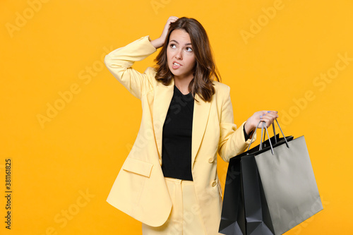 Preoccupied young woman in suit jacket hold package bags with purchases after shopping put hand on head biting lips looking aside up isolated on yellow background studio portrait. Black friday sale.