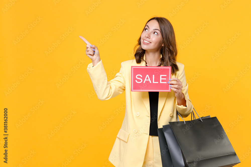 Smiling young woman in basic suit jacket hold package bags with purchases after shopping sign with SALE title pointing index finger aside isolated on yellow background, studio portrait. Black friday. Smiling young woman in basic suit jacket hold package bags with purchases after shopping sign with SALE title pointing index finger aside isolated on yellow background, studio portrait. Black friday.