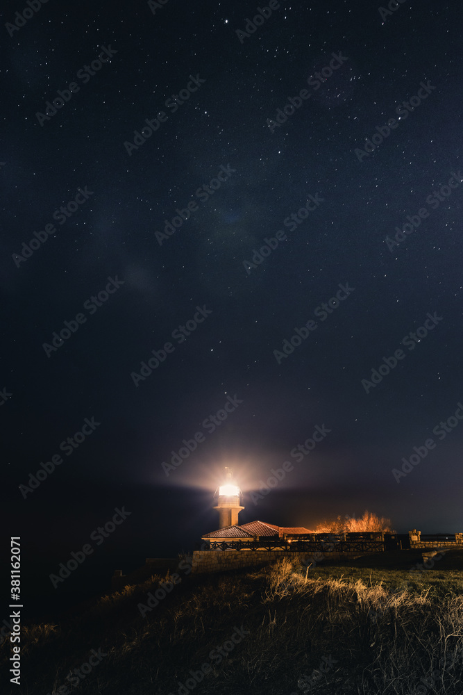 A photography of a lighthouse lighting the night under the stars.
