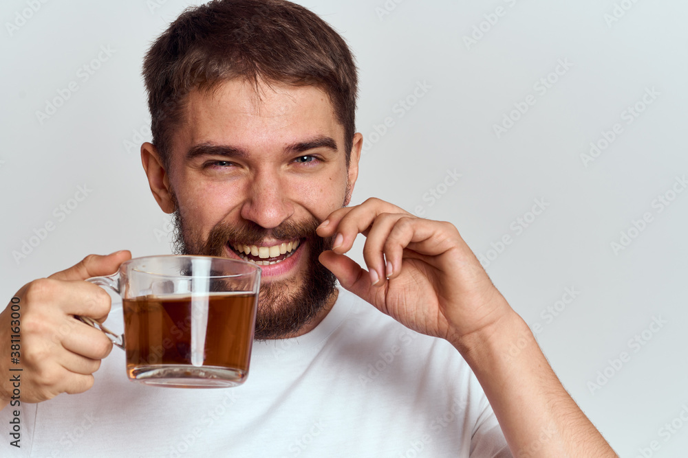 A happy man with a mustache and a beard holds a cup of tea in his hand on a light background and laughs A happy man with a mustache and a beard holds a cup of tea in his hand on a light background and laughs