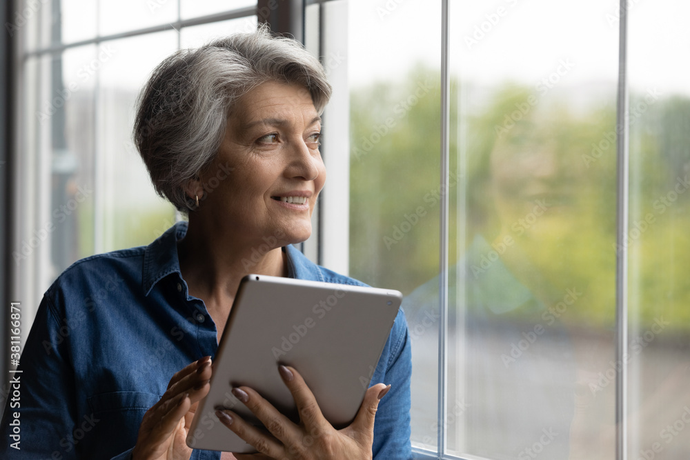 Happy mature grey-haired Caucasian woman use modern tablet gadget look in window distance thinking. Smart senior 60s grandmother browse internet on pad device at home. Elderly and technology concept. Happy mature grey-haired Caucasian woman use modern tablet gadget look in window distance thinking. Smart senior 60s grandmother browse internet on pad device at home. Elderly and technology concept.