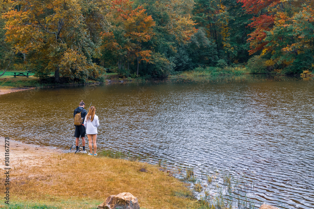 A photograph of two teens fishing from the shore of a lake surrounded by trees showing autumn colors.