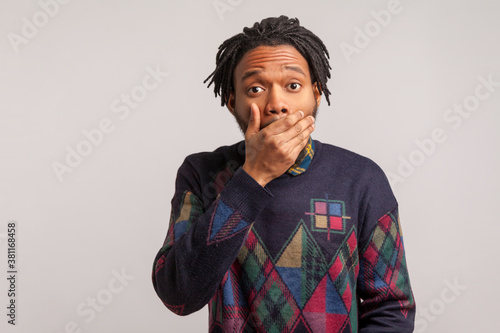 Afraid african man with dreadlock closing mouth with hand looking at camera with big eyes, keeping secret, shocked wondered with news. Indoor studio shot isolated on gray background