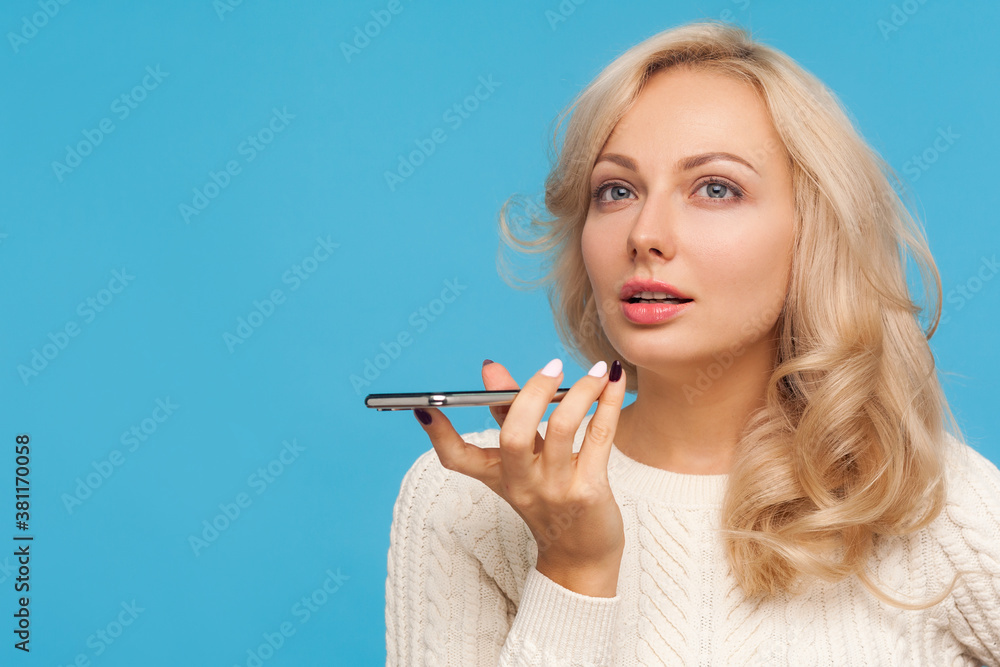 Obraz premium Closeup beautiful woman with curly blond hair holding smartphone in hand dictating note to voice assistant, template. Indoor studio shot isolated on blue background