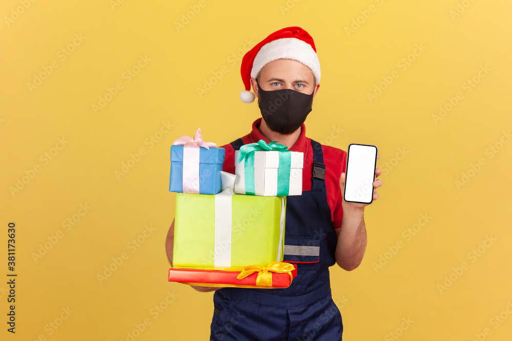 Logistic man in protective mask, uniform and santa claus hat showing christmas presents and smartphone, easy order and delivery during quarantine. Indoor studio shot isolated on yellow background Logistic man in protective mask, uniform and santa claus hat showing christmas presents and smartphone, easy order and delivery during quarantine. Indoor studio shot isolated on yellow background