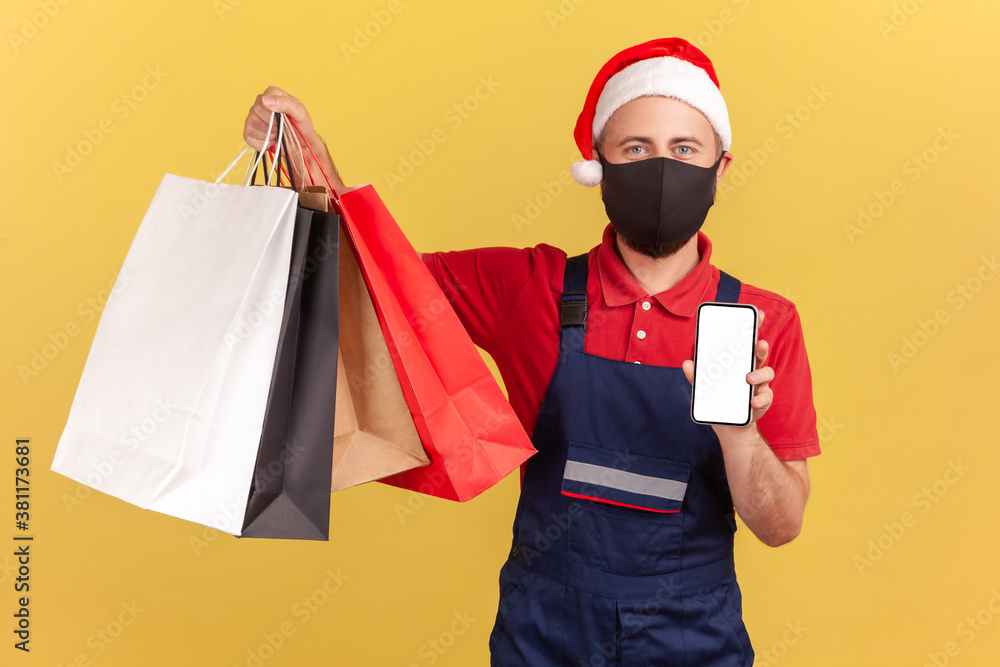 Male courier in protective medical mask and santa claus red hat holding shopping bags and smartphone, easy order and delivery app, quarantine services. Indoor studio shot isolated on yellow background