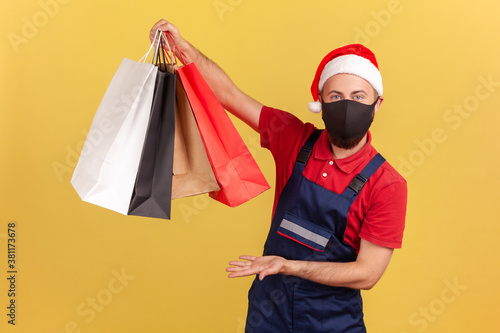 Deliveryman in santa claus hat and black protective face mask showing shopping bags, easy goods order and delivery on quarantine. Indoor studio shot isolated on yellow background
