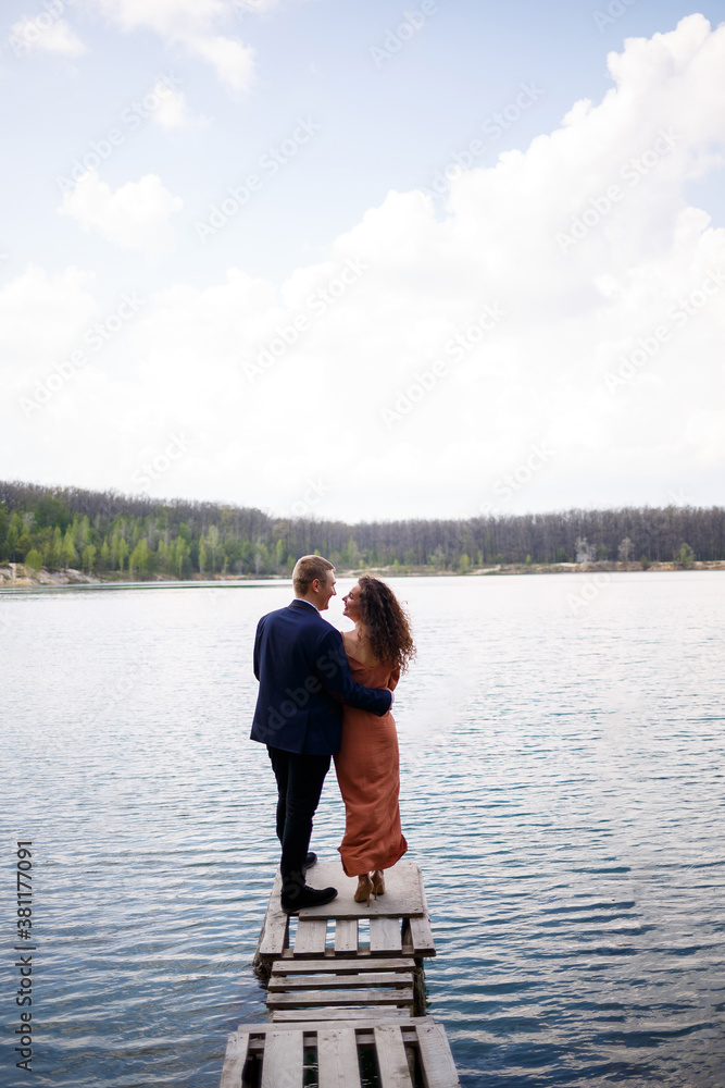A young couple holding hands on a wooden bridge in the middle of a blue lake. Masonry on the island on a background of trees. Nature, landscape. Romance and love, happy couple