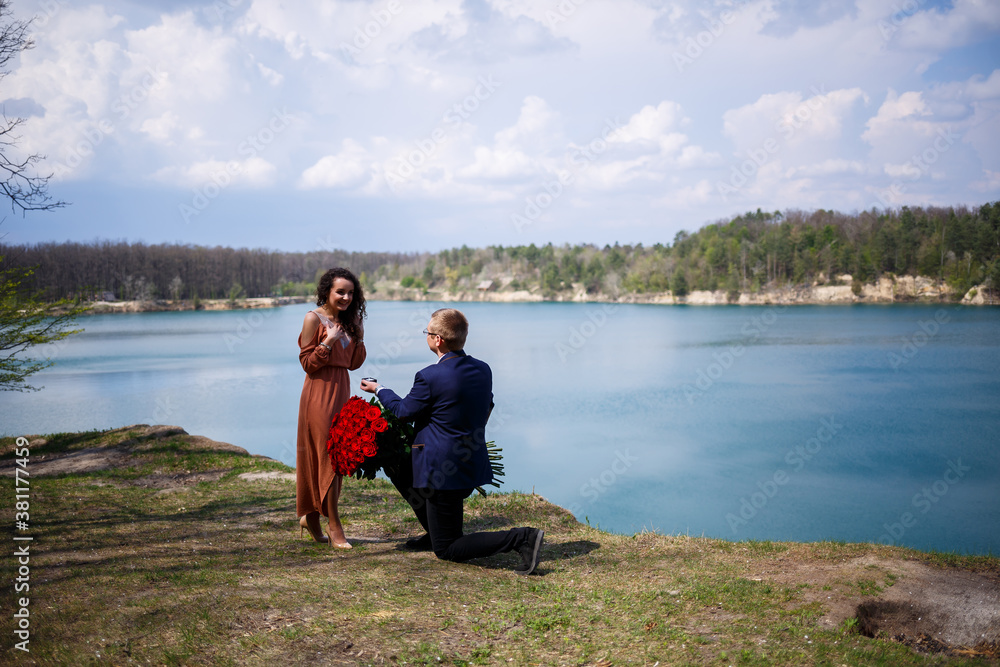 Wedding background in the forest: a man made a surprise, a ring gives a beautiful girl a declaration of love and a marriage proposal. Girl with a smile on her face and a bouquet of red roses