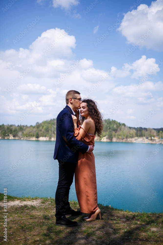 Happy and young newlyweds go holding hands and laughing, on the background of a lake and a green meadow. Groom and beautiful bride with curly hair are walking in the meadow. Joyful summer day Happy and young newlyweds go holding hands and laughing, on the background of a lake and a green meadow. Groom and beautiful bride with curly hair are walking in the meadow. Joyful summer day