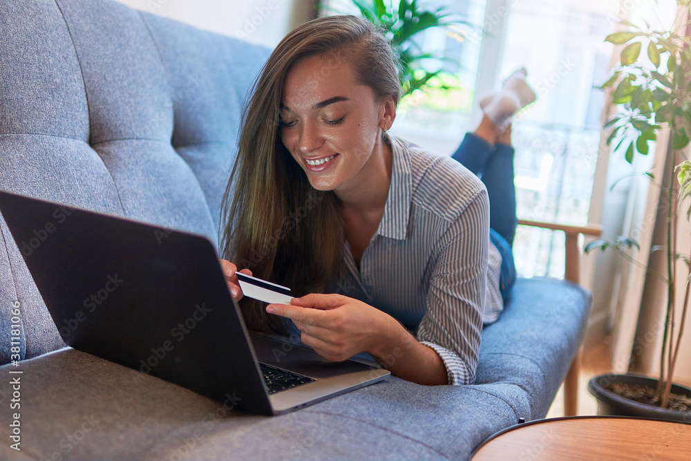 Naklejka premium Happy smiling young casual woman lying on a sofa, resting and shopping online at a computer at cozy comfy home