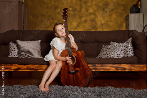 Pretty young four-year-old girl with guitar sitting on sofa in interior of room. Happy child at home. Concept of house learning or playing guitar at home. Copyright space for site or banner