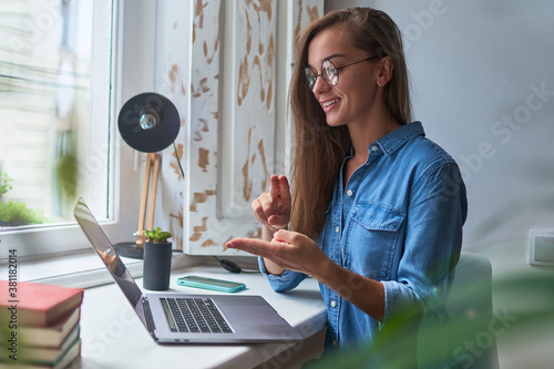 Happy young smiling casual woman learning and communicates in sign language online at a laptop at cozy comfy home by the window