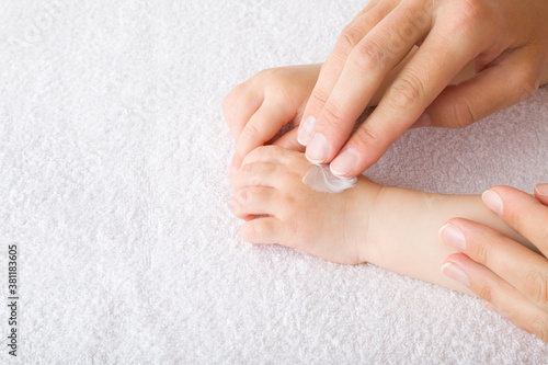 Young mother fingers applying moisturizing cream on baby hand on white towel. Care about children clean and soft body skin. Side view. Closeup.