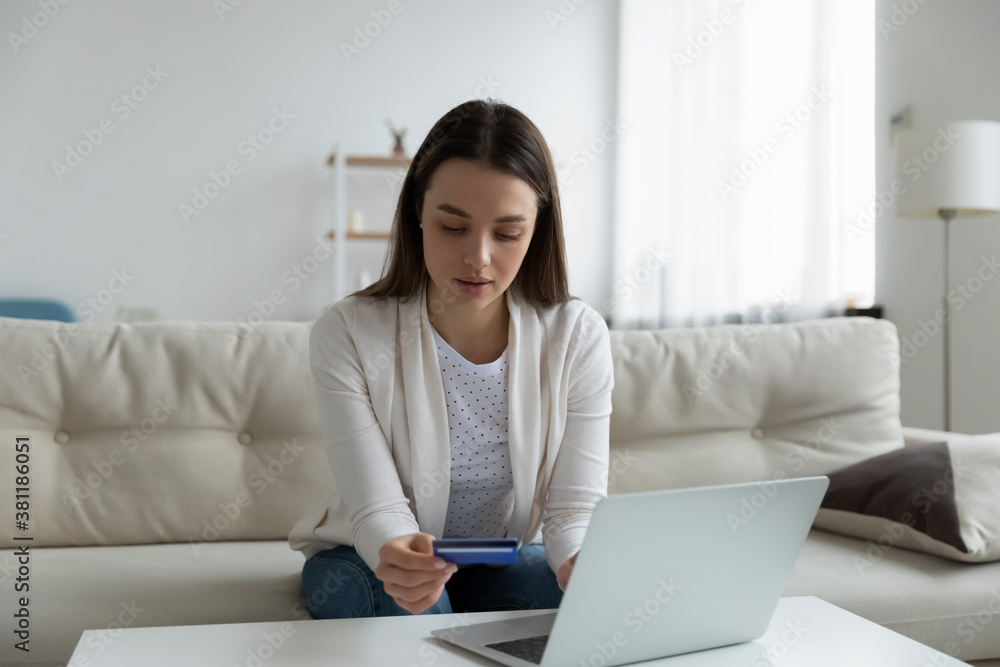 Young Caucasian woman sit on sofa in living room shopping online on laptop with credit card. Millennial female make internet payment on computer, enter bank details, use secure web service system. Young Caucasian woman sit on sofa in living room shopping online on laptop with credit card. Millennial female make internet payment on computer, enter bank details, use secure web service system.