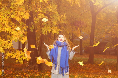 Outdoors lifestyle fashion image of happy beautiful girl throwing leaves up in the air in autumn park. With arms raised up. Wearing stylish grey coat and blue scarf. Happiness carefree.
