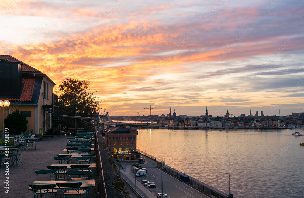 Beautiful sunset view in Stockholm. Colorful golden orange pink sky over the capital of Sweden. Embankment with outdoors cafe. Gamla Stan Old Town embankment with silhouettes of buildings and towers.