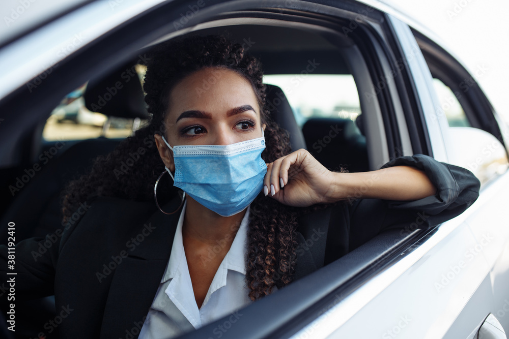 Young business woman fixing and adjusting her medical mask while sitiing in the car behind the steering wheel. Business trips during pandemic, new normal and coronavirus travel safety concept. Young business woman fixing and adjusting her medical mask while sitiing in the car behind the steering wheel. Business trips during pandemic, new normal and coronavirus travel safety concept.