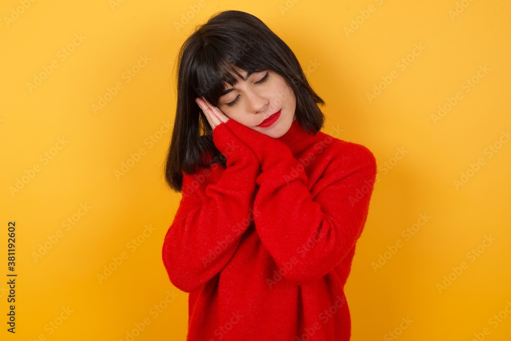 Caucasian brunette woman wearing red casual sweater isolated over yellow background sleeping tired dreaming and posing with hands together while smiling with closed eyes. Caucasian brunette woman wearing red casual sweater isolated over yellow background sleeping tired dreaming and posing with hands together while smiling with closed eyes.